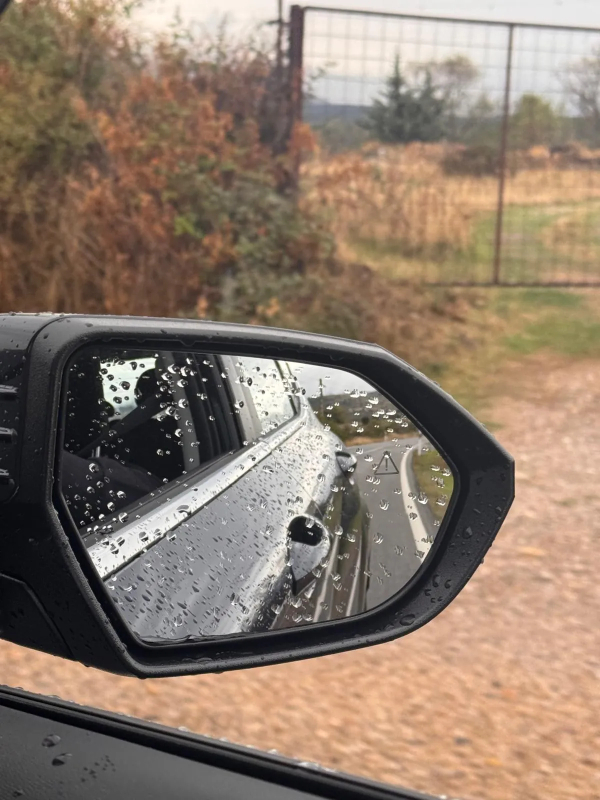 Retrovisor exterior del Hyundai KONA cubierto de gotas de lluvia reflejando una carretera con curvas