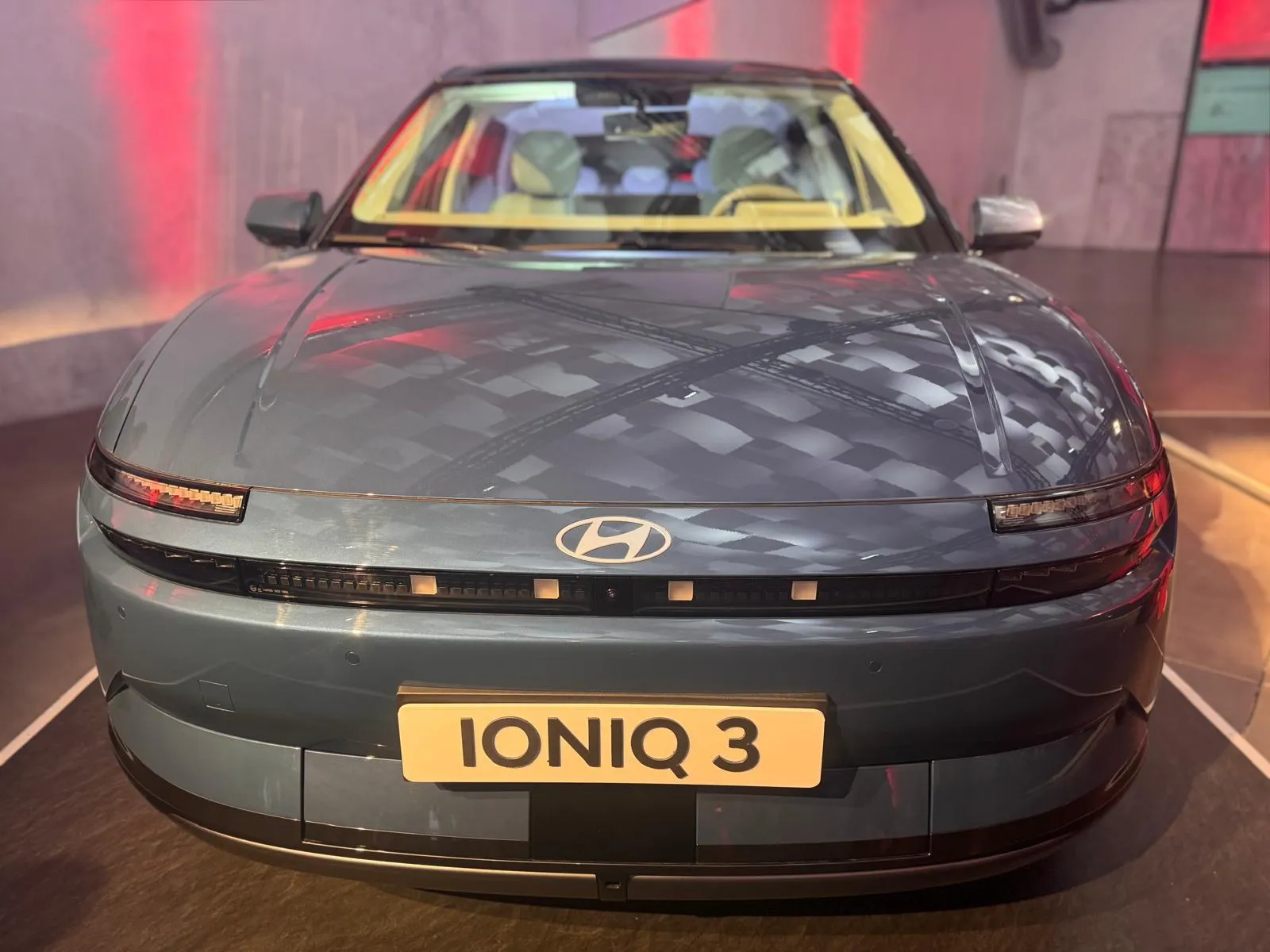 Frontal del Hyundai IONIQ 3 en gris azulado con la franja de luces de píxeles y el logotipo de Hyundai en el capó
